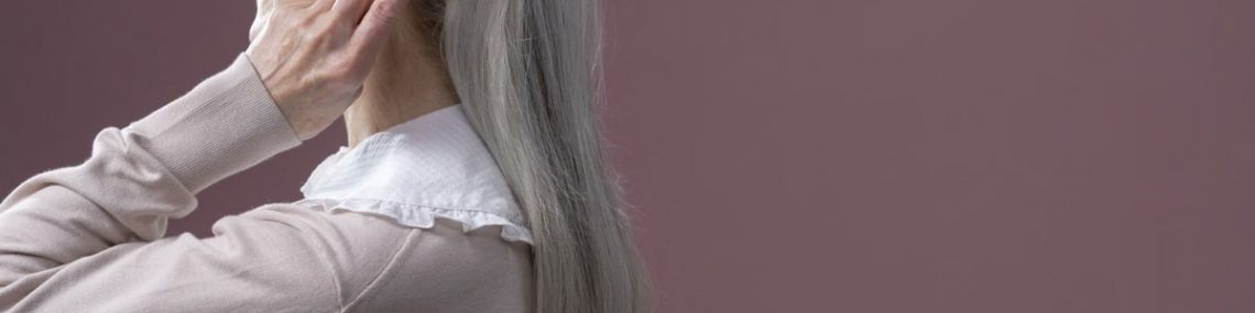Elderly woman with long grey hair combing it, set against a purple background.